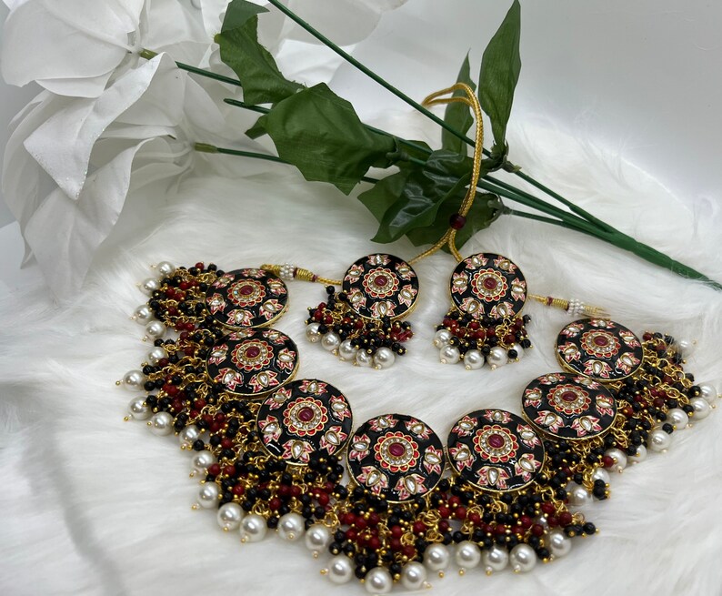 May include: A black and gold necklace with red and pink floral accents. The necklace is adorned with black and white beads and pearls. The necklace is paired with matching earrings.