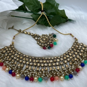 Puede incluir: Un conjunto de collar y maang tikka dorado con un gran colgante adornado. El collar presenta una fila de piedras de forma cuadrada y est&aacute; adornado con peque&ntilde;as perlas blancas y cuentas de colores.