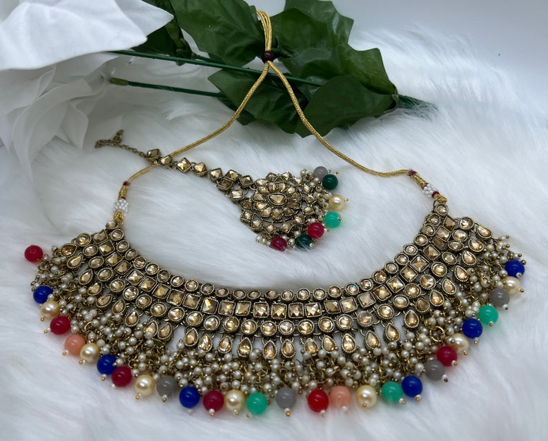 Puede incluir: Un conjunto de collar y maang tikka dorado con un gran colgante adornado. El collar presenta una fila de piedras de forma cuadrada y est&aacute; adornado con peque&ntilde;as perlas blancas y cuentas de colores.