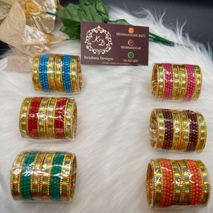 May include: Six sets of gold bangles with colorful beads. Each set has a different color of beads, including blue, red, brown, green, and pink. The bangles are wrapped in plastic and are ready to be shipped.
