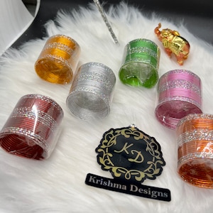 May include: A set of six colorful metal bangles with glitter accents. The bangles are packaged in clear plastic and are arranged on a white fuzzy surface. The bangles are in the colors orange, silver, green, pink, red, and copper.