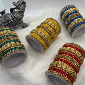 May include: Four sets of bangles with gold and green, gold and yellow, gold and red, and gold and blue accents. The bangles are stacked on top of each other and are displayed on a white surface.