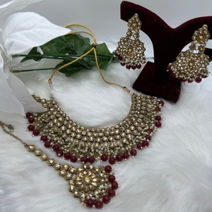 Puede incluir: Un conjunto de collar de novia dorado y burdeos con un collar estilo choker, pendientes a juego y un tikka. El collar y los pendientes presentan dise&ntilde;os intrincados con piedras transparentes y detalles de perlas. El tikka tiene un colgante cuadrado con un dise&ntilde;o floral.