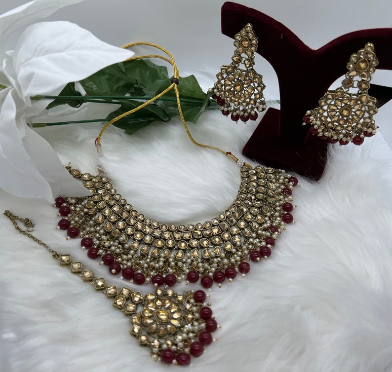 Puede incluir: Un conjunto de collar de novia dorado y burdeos con un collar estilo choker, pendientes a juego y un tikka. El collar y los pendientes presentan dise&ntilde;os intrincados con piedras transparentes y detalles de perlas. El tikka tiene un colgante cuadrado con un dise&ntilde;o floral.