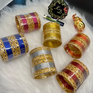 May include: A set of six gold bangle bracelets with decorative designs. The bracelets are in various colors, including red, blue, silver, and gold. The bracelets are stacked on a white fluffy surface.