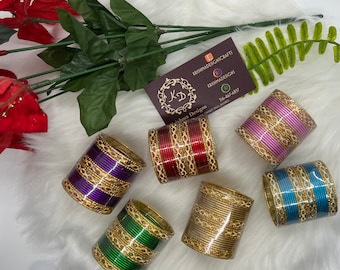 May include: Six different colored bangles with gold accents. The bangles are stacked on a white fluffy surface. The bangles are made of metal and have a shiny finish. The bangles are all different colors, including red, purple, pink, blue, and green.