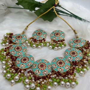 May include: A turquoise and gold necklace set with red and white accents. The necklace features a large round pendant with intricate floral designs and is adorned with white and green beads. The earrings are smaller versions of the pendant.