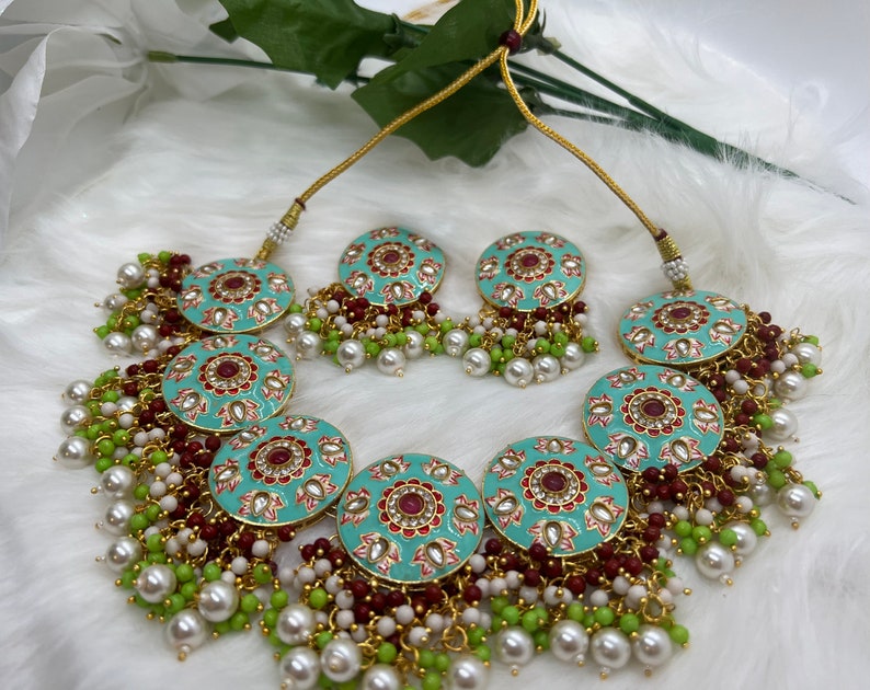 May include: A turquoise and gold necklace set with red and white accents. The necklace features a large round pendant with intricate floral designs and is adorned with white and green beads. The earrings are smaller versions of the pendant.