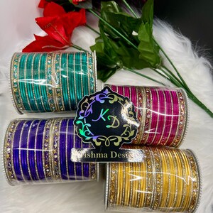 Op de afbeelding: Vier sets armbanden in verschillende kleuren, waaronder turquoise, roze, paars en goud. Elke set heeft een zilveren band met strass. De armbanden zijn op elkaar gestapeld en bevinden zich in een doorzichtige plastic container. Het label "Krishna Designs" is zichtbaar op de afbeelding.