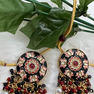 May include: A pair of black and gold earrings with red and white enamel detailing. The earrings feature a round pendant with a floral design and a dangling chain of black and red beads.
