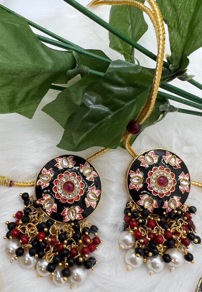 May include: A pair of black and gold earrings with red and white enamel detailing. The earrings feature a round pendant with a floral design and a dangling chain of black and red beads.
