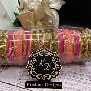 Op de afbeelding: Een set roze en gouden armbanden met ingewikkelde ontwerpen. De armbanden zijn in een cirkelvormige opstelling gerangschikt en verpakt in een doorzichtige plastic container. De container heeft een label met de tekst "Krishna Designs".