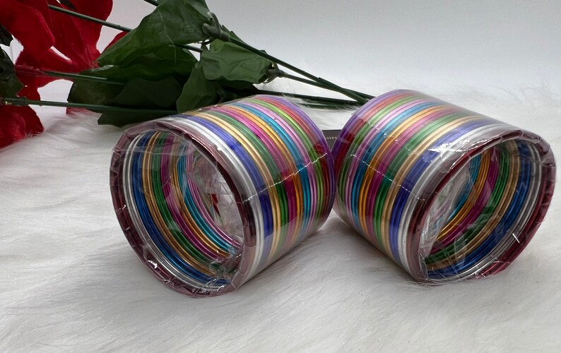 May include: A set of colorful metal bangles in a plastic package. The bangles are arranged in a circle and are all different colors, including red, orange, yellow, green, blue, purple, and silver. The bangles are shiny and have a metallic finish.