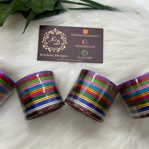 May include: Four stacks of colorful bangles, each stack containing approximately 20 bangles. The bangles are made of metal and have a shiny finish. The bangles are arranged in a circle, with the stacks of bangles facing the camera. The stacks of bangles are on a white fuzzy surface.