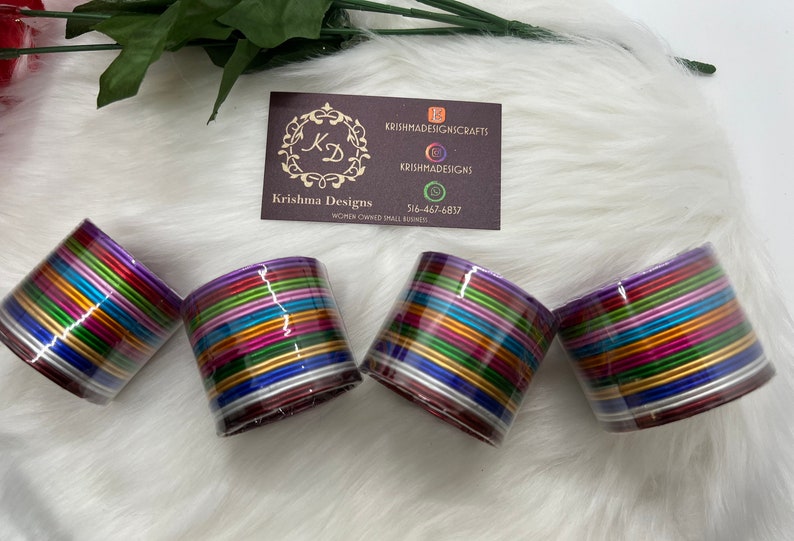 May include: Four stacks of colorful bangles, each stack containing approximately 20 bangles. The bangles are made of metal and have a shiny finish. The bangles are arranged in a circle, with the stacks of bangles facing the camera. The stacks of bangles are on a white fuzzy surface.