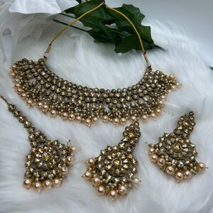 Puede incluir: Un conjunto de collar y pendientes de tono dorado con piedras transparentes y detalles de perlas. El collar es de estilo gargantilla con un dise&ntilde;o en capas. Los pendientes son de estilo colgante con forma redonda.