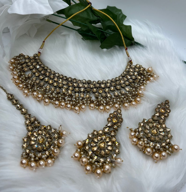 Puede incluir: Un conjunto de collar y pendientes de tono dorado con piedras transparentes y detalles de perlas. El collar es de estilo gargantilla con un dise&ntilde;o en capas. Los pendientes son de estilo colgante con forma redonda.