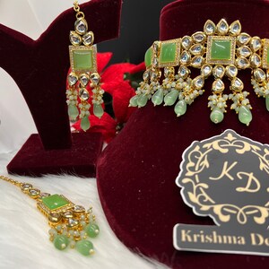 May include: A green and gold Indian bridal jewelry set with a choker necklace, earrings, and a maang tikka. The set features green stones and white pearls.