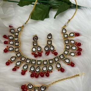 Peut inclure: Un ensemble de collier en or avec des perles rouges et vertes, présentant un motif en forme de larme et de carré avec des pierres blanches. L'ensemble comprend un collier, des boucles d'oreilles et un maang tikka.