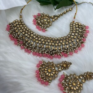 Puede incluir: Un conjunto de collar y pendientes de novia en oro y rosa con un gran colgante adornado y pendientes a juego. El collar presenta un dise&ntilde;o en capas con cuentas rosas y perlas. Los pendientes tambi&eacute;n est&aacute;n adornados con cuentas rosas y perlas.