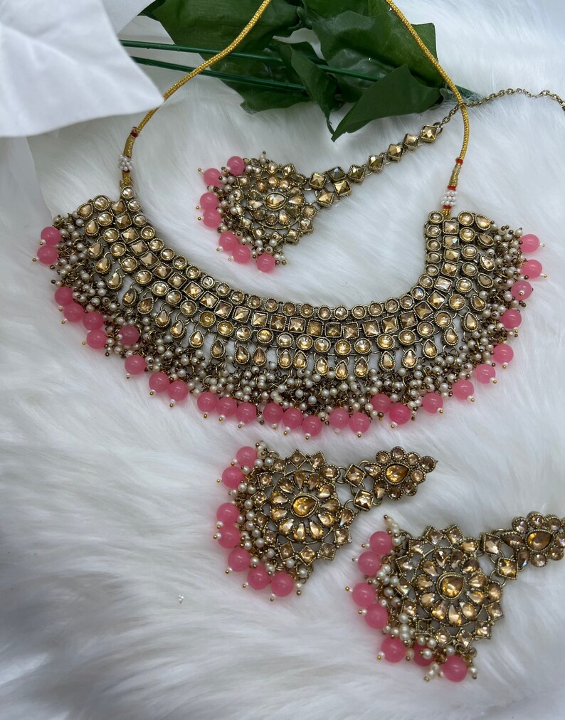 Puede incluir: Un conjunto de collar y pendientes de novia en oro y rosa con un gran colgante adornado y pendientes a juego. El collar presenta un dise&ntilde;o en capas con cuentas rosas y perlas. Los pendientes tambi&eacute;n est&aacute;n adornados con cuentas rosas y perlas.