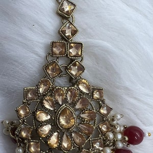 Puede incluir: Tocado dorado con un gran dise&ntilde;o circular adornado con brillantes transparentes. El tocado est&aacute; acentuado con una cadena de brillantes cuadrados m&aacute;s peque&ntilde;os y una franja de perlas blancas y cuentas burdeos.