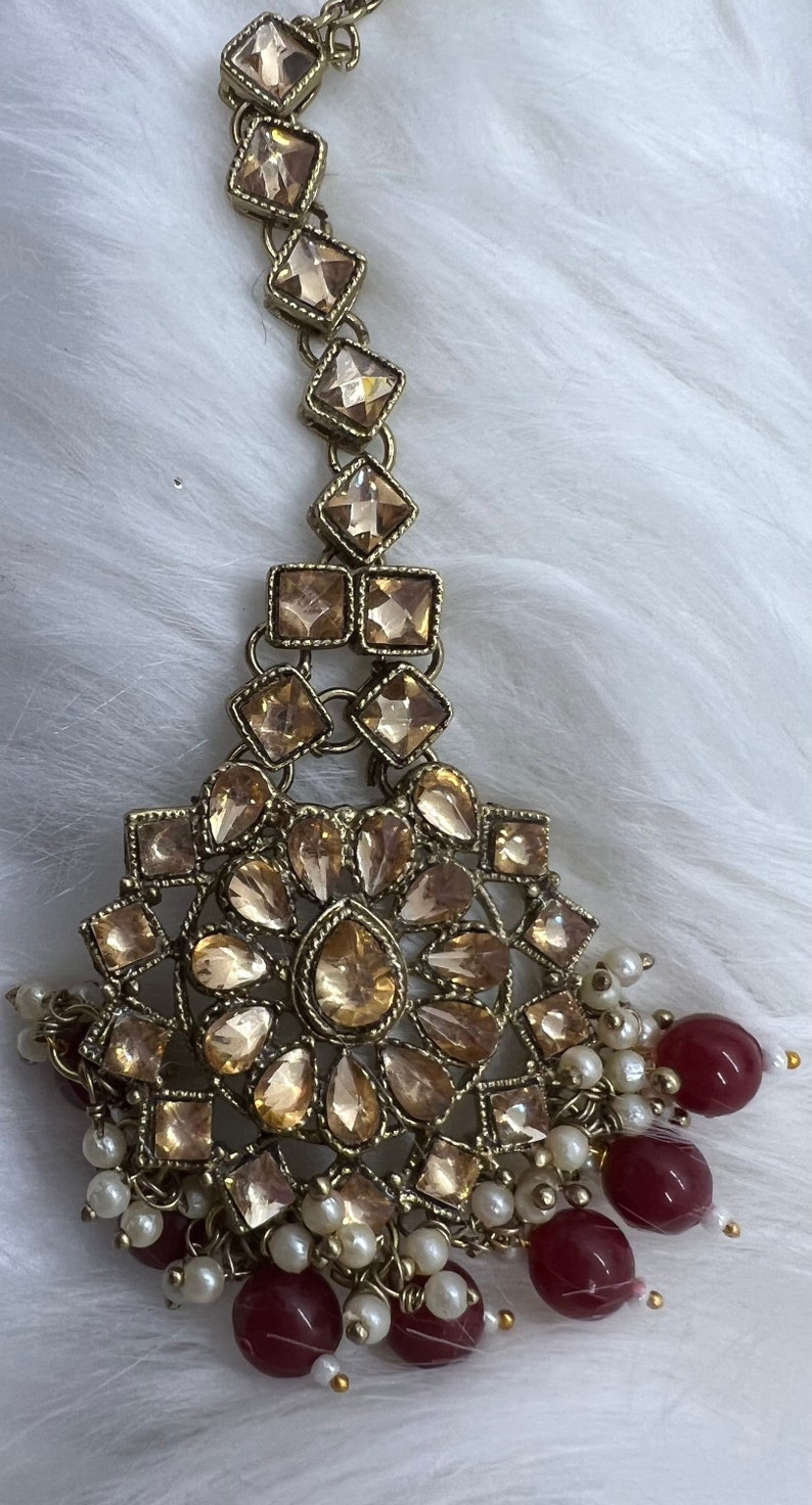 Puede incluir: Tocado dorado con un gran dise&ntilde;o circular adornado con brillantes transparentes. El tocado est&aacute; acentuado con una cadena de brillantes cuadrados m&aacute;s peque&ntilde;os y una franja de perlas blancas y cuentas burdeos.