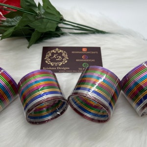 May include: A set of colorful metal bangles, packaged in clear plastic. The bangles are arranged in a stack, with each bangle a different color. The colors include purple, blue, green, yellow, orange, and red. The bangles are all the same size and have a shiny finish.