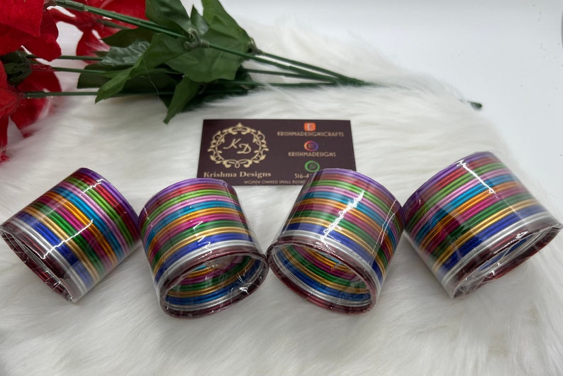 May include: A set of colorful metal bangles, packaged in clear plastic. The bangles are arranged in a stack, with each bangle a different color. The colors include purple, blue, green, yellow, orange, and red. The bangles are all the same size and have a shiny finish.
