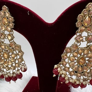 Puede incluir: Un par de pendientes de tono dorado con un dise&ntilde;o en forma de l&aacute;grima. Los pendientes est&aacute;n adornados con diamantes de imitaci&oacute;n transparentes y &aacute;mbar y tienen una cadena colgante de cuentas rojas y blancas.