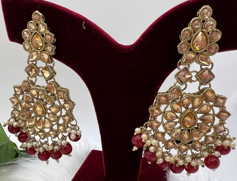 Puede incluir: Un par de pendientes de tono dorado con un dise&ntilde;o en forma de l&aacute;grima. Los pendientes est&aacute;n adornados con diamantes de imitaci&oacute;n transparentes y &aacute;mbar y tienen una cadena colgante de cuentas rojas y blancas.