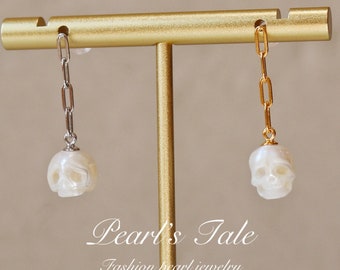 May include: A pair of pearl skull earrings with a gold chain on a gold stand. The earrings are white and have a skull design. The text "Pearl's Tale Fashion pearl jewelry" is below the earrings.