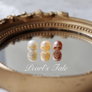 May include: Three small skull-shaped pendants made of white, cream, and reddish-brown pearl. The pendants are displayed on a mirrored surface with the text "Pearl's Tale Fashion Pearl Jewelry".