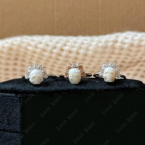 May include: Three rings featuring skull-shaped centrepieces. The rings have silver or gold bands, with a skull design and a crown-like embellishment. The skulls are white, and the crowns are adorned with clear or rose-coloured stones. The rings are displayed on a black surface.
