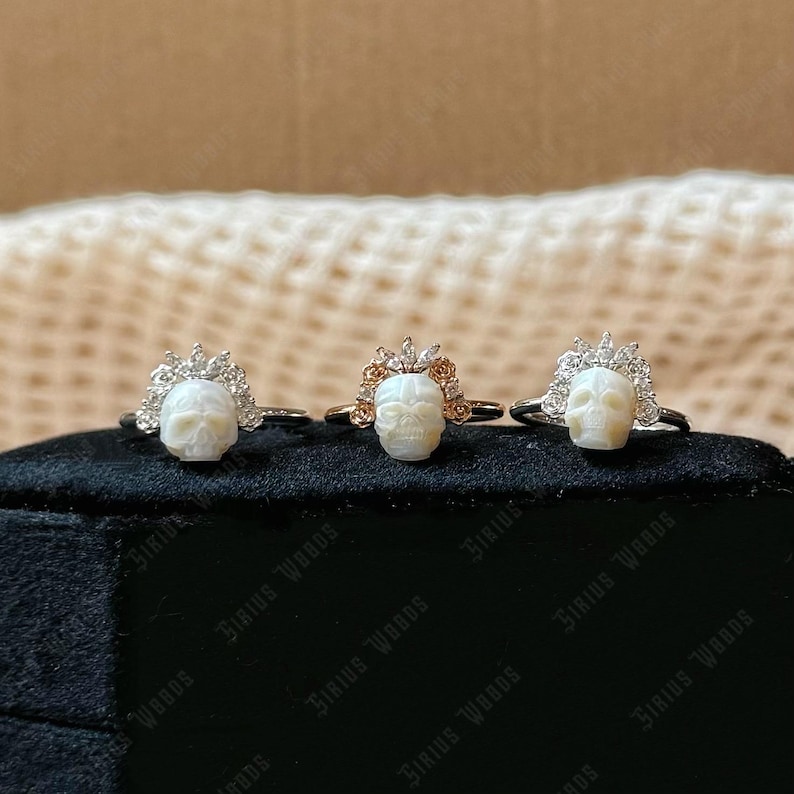 May include: Three rings featuring skull-shaped centrepieces. The rings have silver or gold bands, with a skull design and a crown-like embellishment. The skulls are white, and the crowns are adorned with clear or rose-coloured stones. The rings are displayed on a black surface.