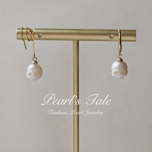 May include: A pair of gold skull earrings with white pearl accents. The earrings are hanging from a gold stand. The text "Pearl's Tale Fashion Pearl Jewelry" is visible below the earrings.