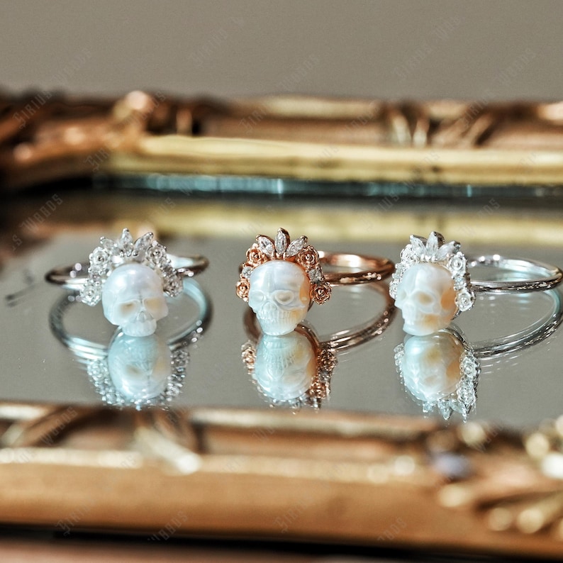 May include: Three rings with skull designs. One ring has a silver band, a skull, and a halo of small stones. Another ring features a rose gold band, a skull, and a crown of small stones. The third ring has a silver band with a skull and a halo of small stones.