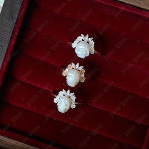 May include: Three skull-shaped rings with intricate detailing. Each ring features a carved skull, surrounded by a halo of small, sparkling stones. The rings are displayed on a deep red velvet surface.