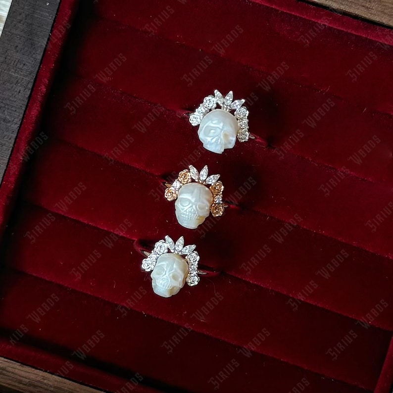 May include: Three rings with skull-shaped designs. Each ring has a carved skull, encircled by a halo of small, sparkling stones. The rings are presented on a deep red velvet surface.