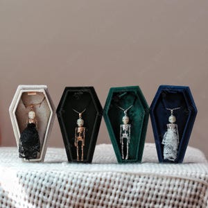 May include: Four skeleton pendant necklaces presented in coffin-shaped boxes. The necklaces feature detailed skeleton charms with different outfits, including a black lace dress and a white lace dress. The boxes are in grey, black, green, and blue.
