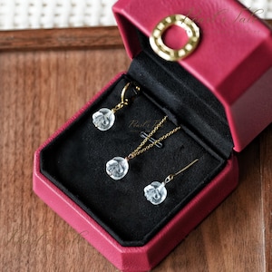 Puede incluir: Un conjunto de joyas que incluye un collar, pendientes y un colgante, presentado en una caja de joyería roja. El conjunto incluye cadenas y detalles dorados, con piedras preciosas transparentes en forma de rosa. La caja está abierta, revelando las joyas.