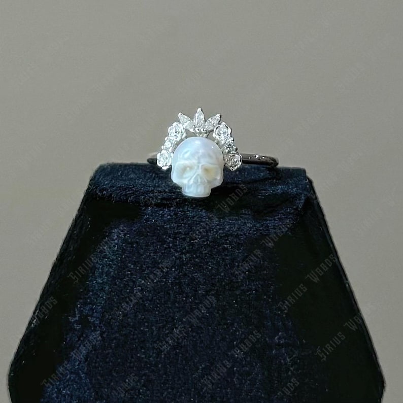 May include: A silver ring featuring a carved skull with a floral crown. The skull is white, and the crown is adorned with small, sparkling accents. The ring is displayed on a black velvet surface.