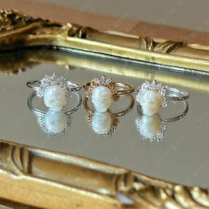 May include: Three rings featuring skull pendants. The rings have silver and gold bands, with the skull pendants in the centre. Each skull is encircled by a decorative halo of crystals or small flowers. The rings are displayed on a reflective surface.