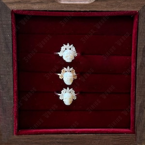 May include: Three skull rings presented in a wooden box with a red velvet lining. Each ring showcases a white skull design, adorned with a crown. The rings are set on silver bands, with one ring featuring gold detailing.