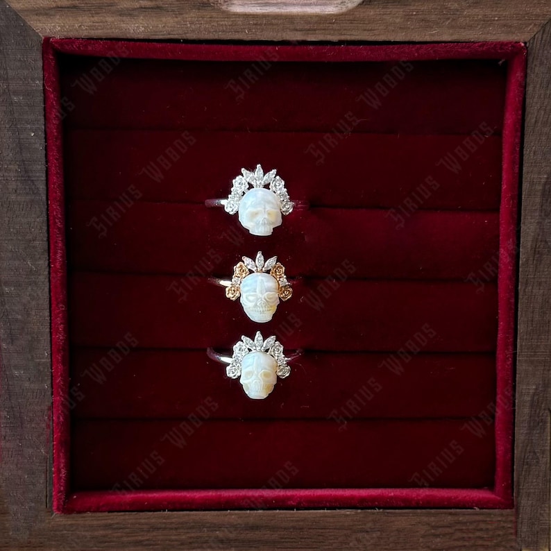 May include: Three skull rings presented in a wooden box with a red velvet lining. Each ring showcases a white skull design, adorned with a crown. The rings are set on silver bands, with one ring featuring gold detailing.