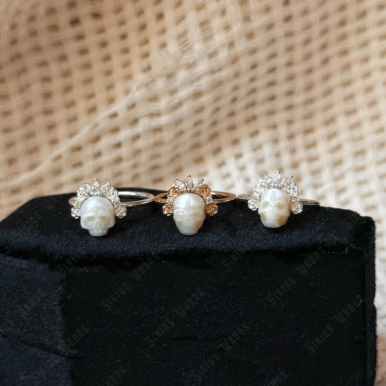 May include: Three skull rings with decorative crowns are presented on a black surface. The rings feature silver and gold bands, and the skulls appear to be carved from a light material. The crowns are detailed with small, sparkling embellishments.