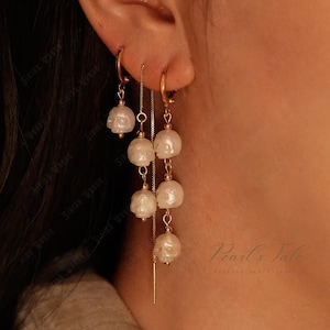 Pode incluir: Brincos dourados com pequenos pingentes de caveira brancos. Um par é um design de argola simples, o outro tem uma corrente com vários pingentes de caveira. Os brincos são exibidos em um fundo quente e neutro.