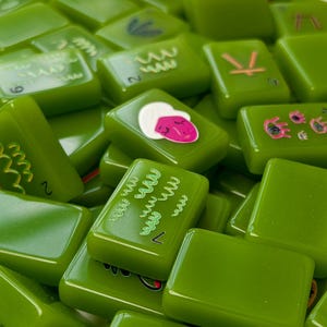May include: A collection of rectangular, glossy olive green tiles featuring various colorful graphic illustrations, including stylized faces, abstract patterns, and symbols. Each tile has a smooth, polished surface and rounded edges.