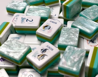May include: A collection of rectangular mahjong tiles featuring a pearlescent light blue top surface, with sides layered in white, green, and teal. The tiles display various printed symbols, numbers, and the word JOKER with fish illustrations.