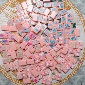May include: A collection of rectangular pink mahjong tiles scattered across a surface. Each tile features engraved designs in blue, green, and pink, including numbers, directional symbols, floral patterns, and joker icons with intricate line work.